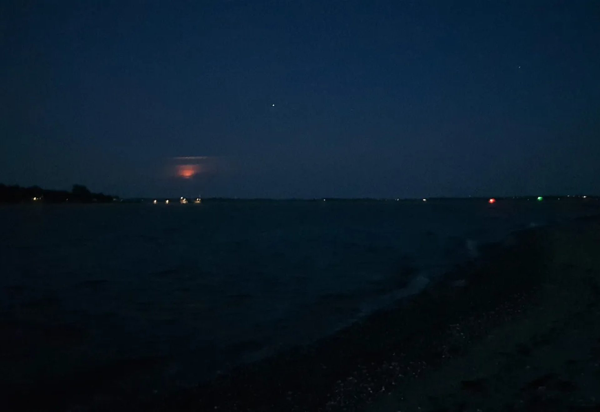 Dunkler Strand bei Nacht mit schwachem Licht am Horizont und wenigen Lichtern in der Ferne.