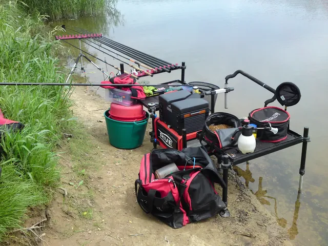 Ein Angelplatz am Ufer mit mehreren Angelruten, Angeltaschen, Eimern und verschiedenen Ausrüstungsgegenständen, die ordentlich neben dem Wasser aufgestellt sind.