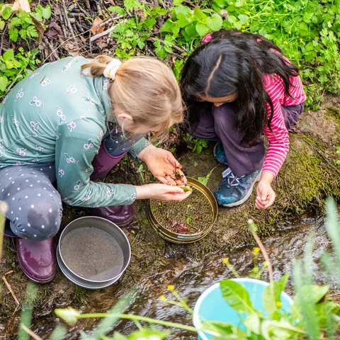 Zwei Kinder sitzen am Ufer eines Bachs und untersuchen mit Sieben Steine oder Erde im Wasser.