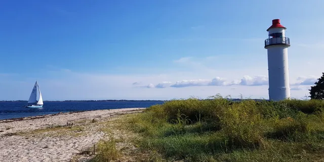 Ein weißer Leuchtturm steht am Rand einer sandigen Küste, links segelt ein Boot auf dem blauen Meer.
