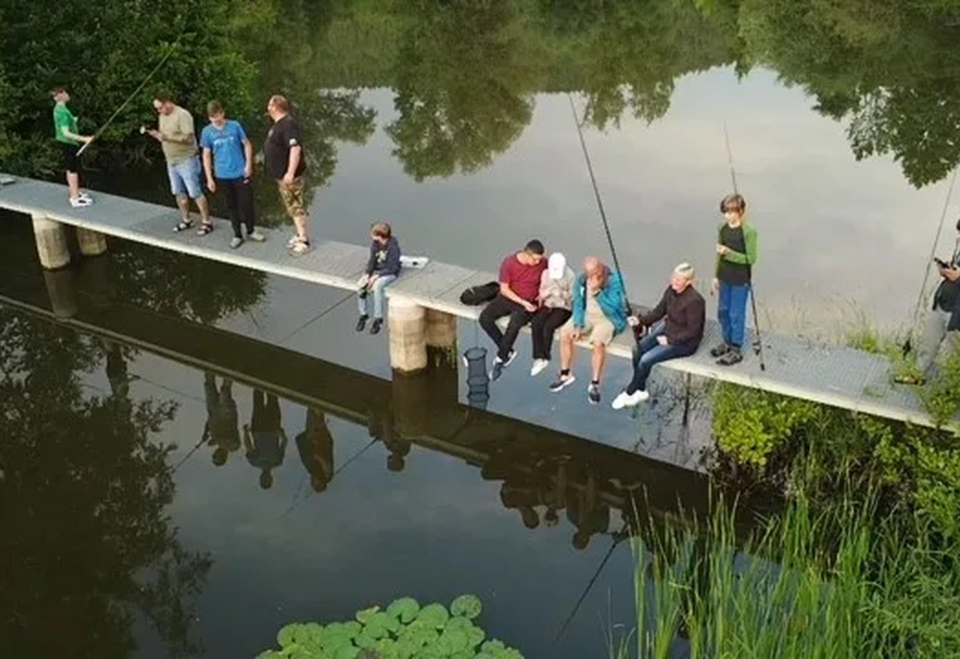 Mehrere Personen sitzen und stehen mit Angelruten auf einem Steg über einem ruhigen, von Bäumen umgebenen See.