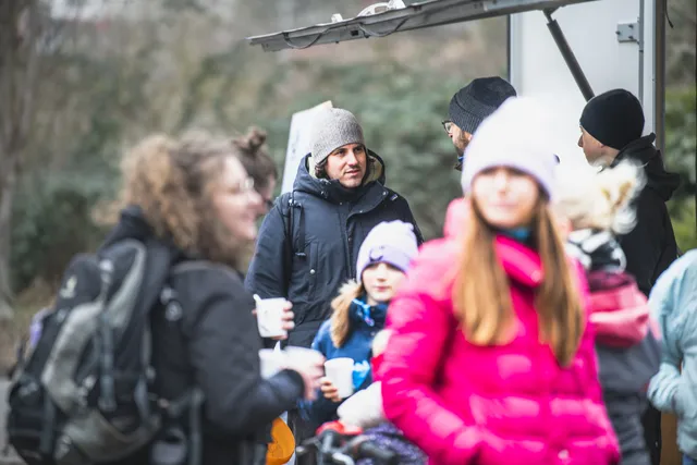 Mehrere Menschen stehen draußen zusammen, einige tragen Winterkleidung und Mützen. Im Vordergrund sind Erwachsene und Kinder zu sehen, von denen manche Becher in der Hand halten. Die Stimmung wirkt winterlich und entspannt.