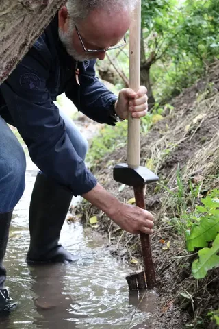 Ein Mann mit Brille gräbt mit einer Schaufel oder Sonde im schlammigen Uferbereich eines kleinen Gewässers.