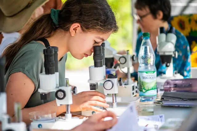 Eine junge Frau betrachtet eine Probe durch ein Mikroskop, weitere Personen arbeiten im Hintergrund an Mikroskopen.