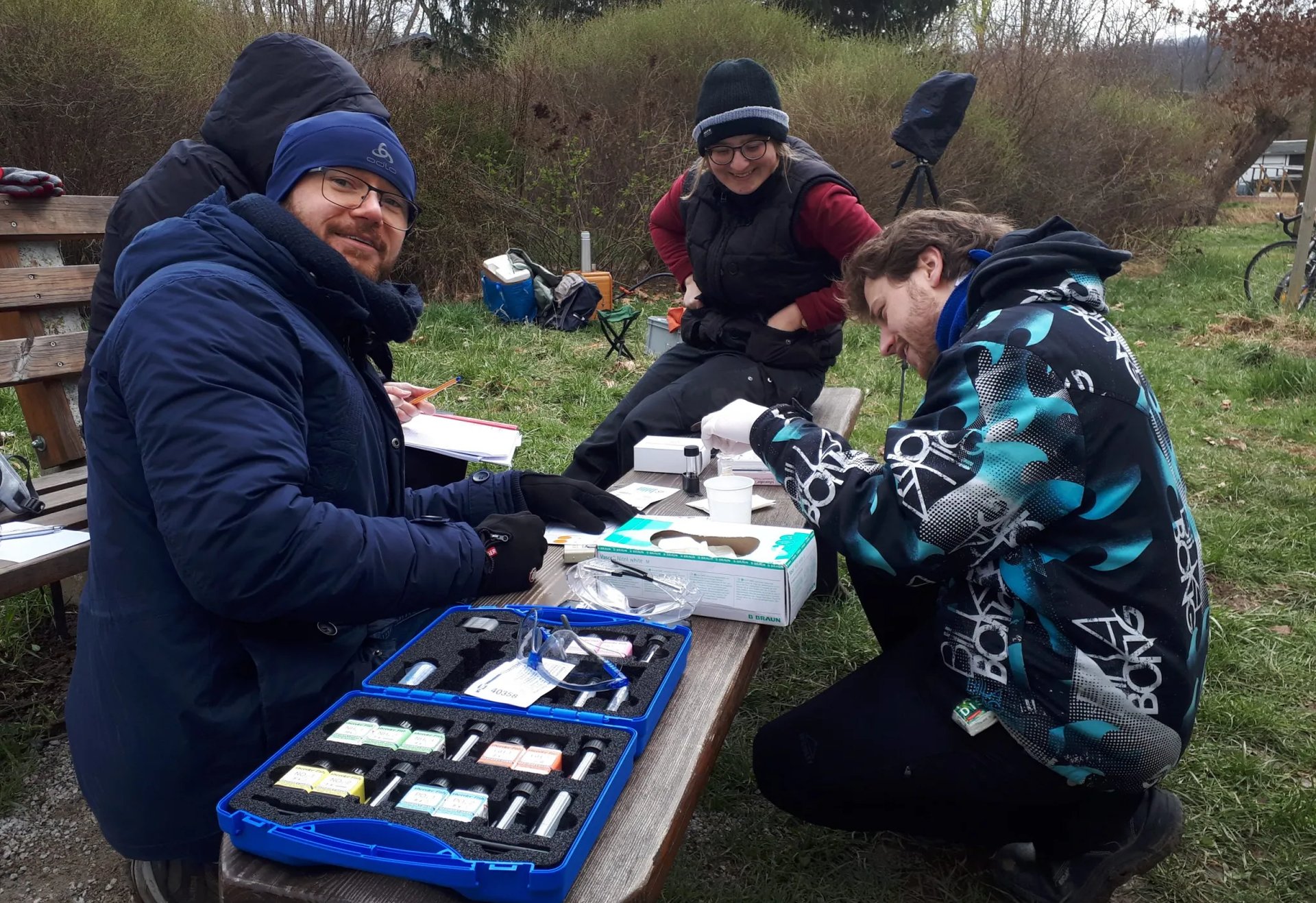 Mehrere Personen sitzen an einem Holztisch im Freien und arbeiten mit einem Mikroskop und Labormaterialien, einige tragen Winterkleidung und Mützen.