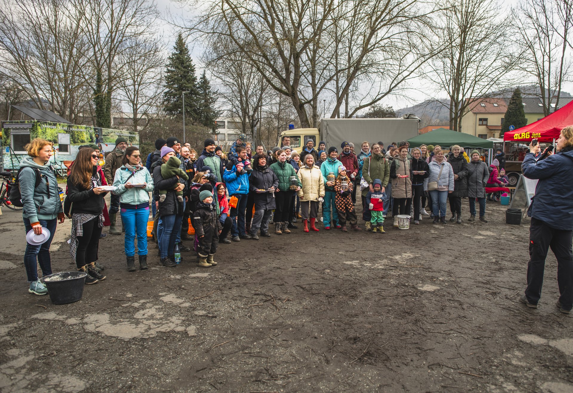 Eine Gruppe von Menschen steht im Freien auf einem matschigen Platz, und posieren um ein Gruppenbild zu machen; im Hintergrund sind Bäume, Fahrzeuge und ein rotes Zelt zu sehen.