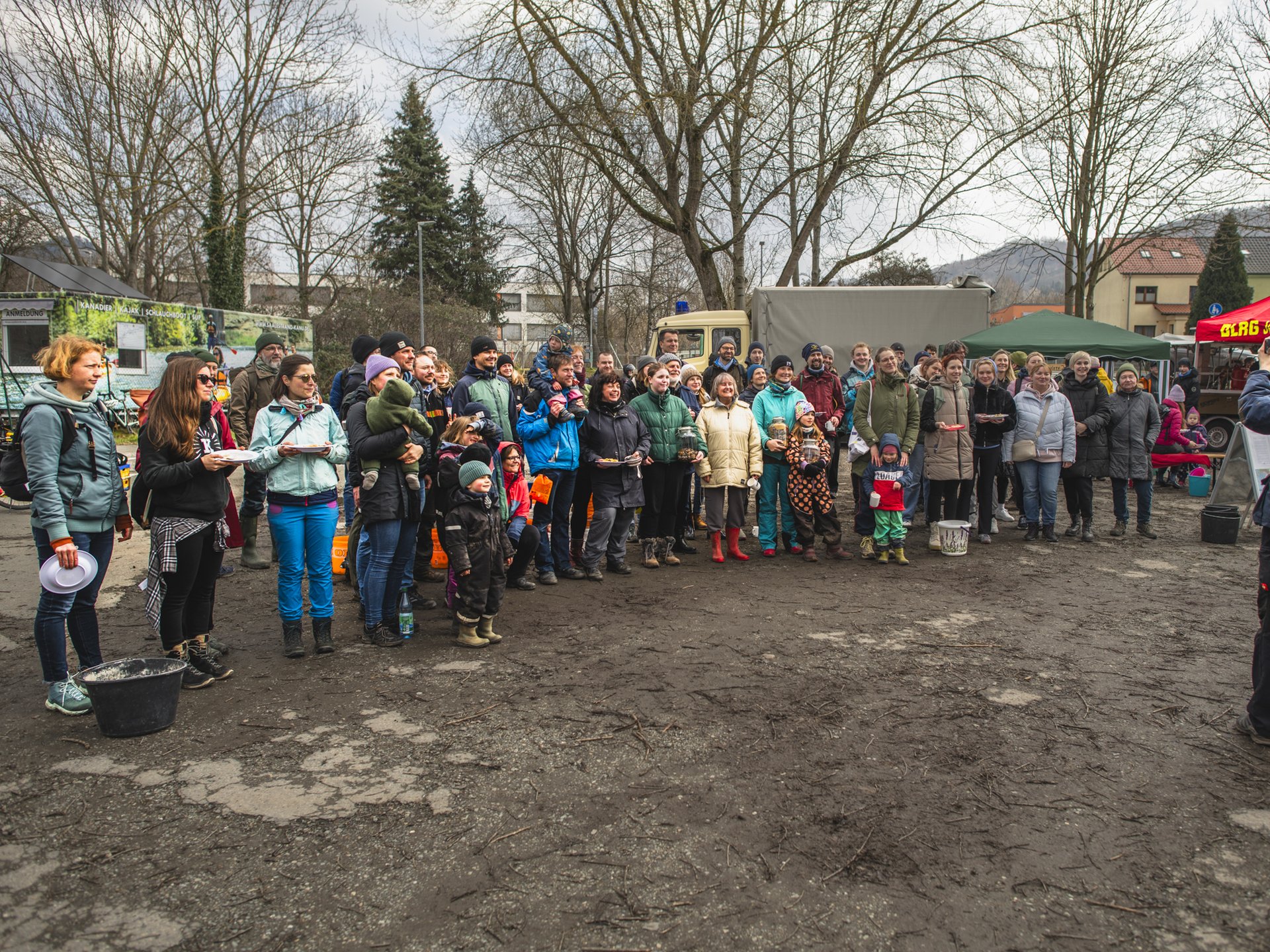 Eine Gruppe von Menschen steht im Freien auf einem matschigen Platz, und posieren um ein Gruppenbild zu machen; im Hintergrund sind Bäume, Fahrzeuge und ein rotes Zelt zu sehen.