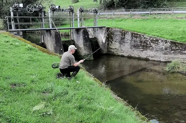 Ein älterer Mann sitzt am Ufer eines kleinen Kanals und angelt neben einer Schleusenanlage.