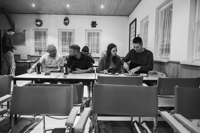 Fünf Menschen sitzen an Tischen in einem Raum und unterhalten sich, während eine Person im Hintergrund steht; der Raum wirkt wie ein Veranstaltungs- oder Versammlungsraum.