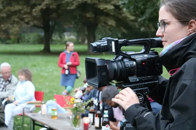 Eine Person filmt mit einer Videokamera im Freien, im Hintergrund sitzen Menschen an einem Tisch im Park.