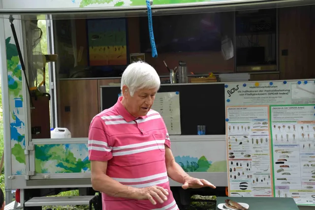 Ein älterer Mann in einem pinken Poloshirt steht vor einem Infostand mit wissenschaftlichen Postern und erklärt etwas.