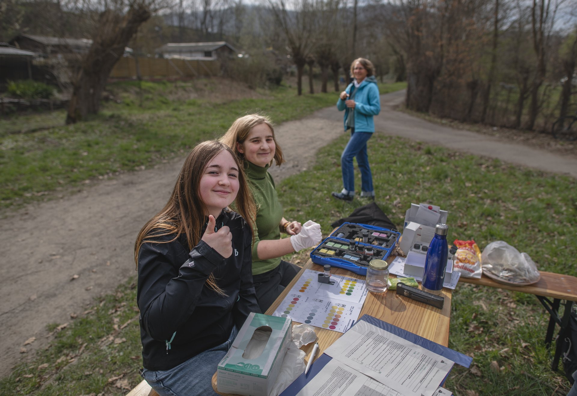 Zwei junge Frauen sitzen an einem Tisch im Freien mit wissenschaftlichen Materialien, eine weitere Person steht im Hintergrund auf einem Weg; alle wirken entspannt und freundlich.