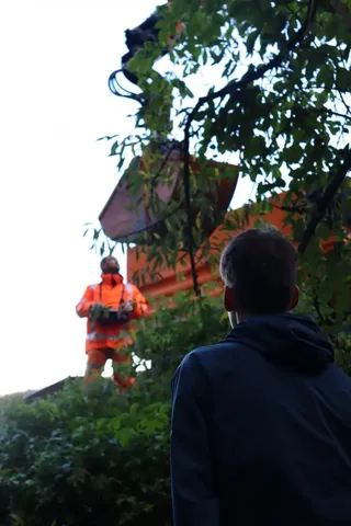 Eine Person in orange Arbeitskleidung steht auf einer erhöhten Fläche hinter Büschen, im Vordergrund blickt eine andere Person im Dunkel gekleidet zu ihr auf.
