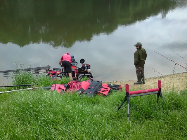 Zwei Personen stehen am Ufer eines ruhigen Sees, umgeben von Angelausrüstung und Ruten.